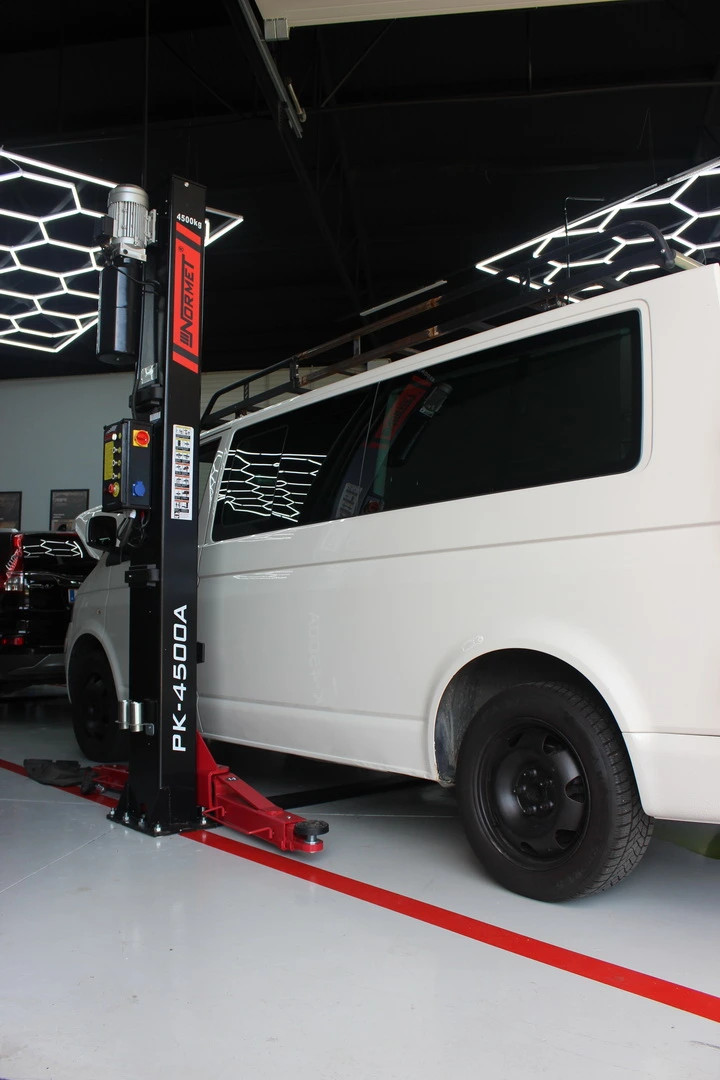 Biele dodávkové auto v dielni s hydraulickým zdvihákom a červenou podperou pri stene.